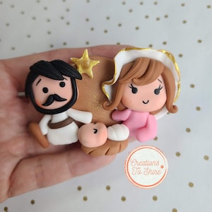 May include: A handmade clay nativity scene featuring a brown-skinned Joseph, a white-skinned Mary, and a baby Jesus in a manger. The figures are stylized with large eyes and simple features. The scene is decorated with gold glitter and a gold star.