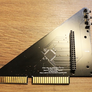 Può includere: Scheda di circuito Amiga nera video e audio RGBtoHDMI CPLD v2.1. Presenta connettori dorati, un processore centrale e diverse porte. Il testo include il numero di versione, informazioni sul copyright e un URL.