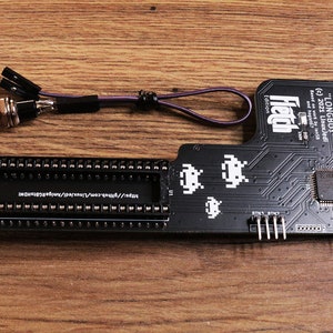 Op de afbeelding: Een zwarte printplaat met het label Fletch Edition Longboy, witte pixelachtige alien-afbeeldingen, een lange pin-header, een geïntegreerde schakeling en een rode drukknop verbonden via een paarse draad met zwarte connectoren.