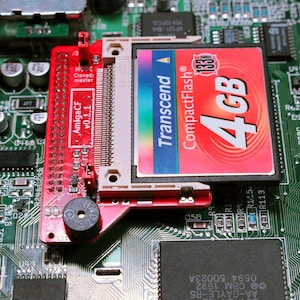 May include: A red AmigaCF v0.1.1 adapter board mounted on a green circuit board, featuring a Transcend 4GB 133x CompactFlash memory card inserted into the slot. The adapter includes a small black circular component and various pins.