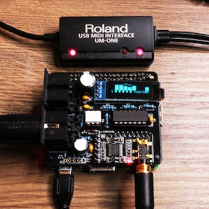 Könnte beinhalten: Ein schwarzes Roland USB MIDI Interface UM-ONE mit roten Kontrollleuchten, verbunden mit einer Leiterplatte mit kleinem Display. Das Display zeigt eine Reihe blauer Balken. Kabel verbinden die Geräte auf einer Holzoberfläche.