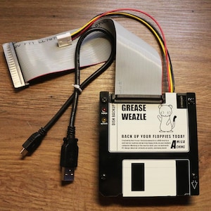 Peut inclure: Un appareil de sauvegarde de disque Grease Weazle noir avec un câble USB et une nappe. L'appareil porte l'inscription "GREASE WEAZLE" et "BACK UP YOUR FLOPPIES TODAY" avec une illustration de belette. L'appareil est sur une surface en bois.