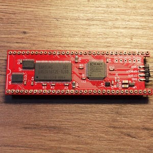 May include: A red circuit board with various integrated circuits and connectors. The board has the text "SAMSUNG 301 K4E6N1612E-TL50". It is designed for electronic components and is placed on a wooden surface.