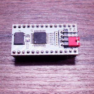 May include: A small, rectangular circuit board with various electronic components. It features a central integrated circuit, a red connector, and multiple pins. The board is light blue and sits on a wooden surface.