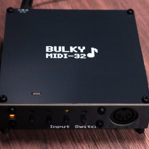 Może przedstawiać: Ciemnoszary, prostokątny kontroler MIDI z białym napisem BULKY MIDI-32 oraz ikoną nuty na górnej powierzchni. Przedni panel zawiera kontrolkę zasilania, kilka małych przełączników oraz okrągłe gniazdo MIDI.
