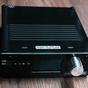 Puede incluir: Dispositivo electrónico negro con una perilla plateada y una pequeña pantalla. El dispositivo tiene una etiqueta que dice "PH541-III FOR C64 FLOPPY DISK DRIVE". La parte superior tiene líneas horizontales y el dispositivo está sobre una superficie de madera.