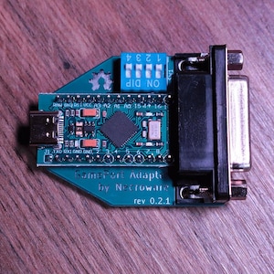 Può includere: Una scheda di circuito GamePort Adapter turchese con un interruttore DIP blu, una porta USB e un connettore argentato. Il testo "GamePort Adapter by Necroware rev 0.2.1" è stampato sulla scheda.
