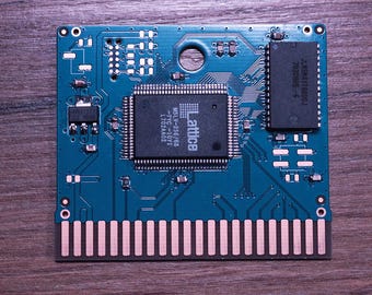 Può includere: Una scheda di circuito turchese con vari componenti elettronici, tra cui un processore centrale etichettato "Lattice" e un chip rettangolare. La scheda ha una serie di pin dorati lungo il bordo inferiore. Lo sfondo è una superficie di legno scuro.