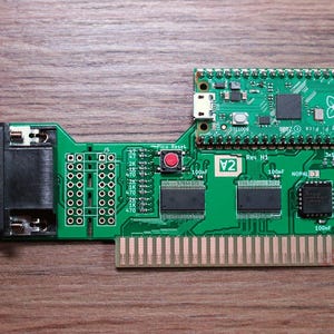 Puede incluir: Una placa de circuito verde con un Raspberry Pi Pico adjunto. La placa tiene varios componentes electrónicos, incluido un botón rojo etiquetado "Pico Reset", y un conector. El texto "V2" es visible.