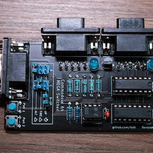 Op de afbeelding: Zwart Commodore 64 MEGA Switcher printplaat met blauwe en zilveren componenten. De printplaat heeft twee zwarte connectoren, blauwe knoppen en de tekst "Commodore 64 MEGA Switcher". De printplaat ligt op een houten oppervlak.