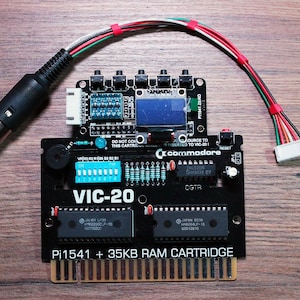 May include: A black VIC-20 cartridge with a Pi1541 + 35KB RAM CARTRIDGE label, featuring a Commodore logo. It has a connected circuit board with a small screen and various components, along with a black connector and a multi-coloured wire.
