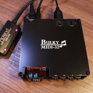 May include: Black Bulky MIDI-32 music interface with a Roland USB MIDI interface connected. The Bulky MIDI-32 has a small display and several input/output jacks. The Roland interface has red indicator lights. The devices are on a wooden surface.