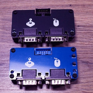 Könnte beinhalten: Zwei elektronische Schnittstellengeräte, eines in Schwarz und eines in Blau, werden gezeigt. Jedes hat ein Joystick- und Maus-Symbol sowie mehrere Anschlüsse und Schrauben. Das blaue Gerät trägt die Aufschriften "JOYSTICK" und "MOUSE".