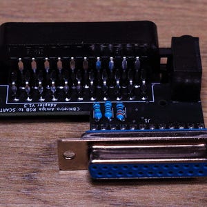 Könnte beinhalten: Ein schwarzer CBMretro Amiga RGB-zu-SCART-Adapter V1.3. Der Adapter hat einen schwarzen SCART-Anschluss, einen blau-silbernen Anschluss und mehrere Pins. Der Adapter liegt auf einer Holzoberfläche.