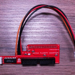 Puede incluir: Un adaptador de disquete Amiga rojo con cables negros, naranjas y rojos y un conector blanco. El adaptador tiene conectores negros y el texto "AMIGA FLOPPY ADAPTER". El dispositivo está sobre una superficie de madera.