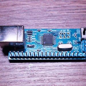 May include: A small, rectangular blue circuit board with various electronic components, including a large central chip, a silver connector, and multiple pins along one edge. The board is set against a wooden surface.