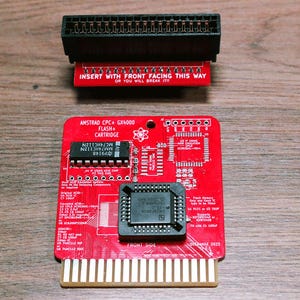 May include: Red Amstrad CPC+ GX4000 Flash+ Cartridge with text "INSERT WITH FRONT FACING THIS WAY" and "OR YOU WILL BREAK IT!". Features a black connector and various electronic components. The cartridge has gold-colored pins.
