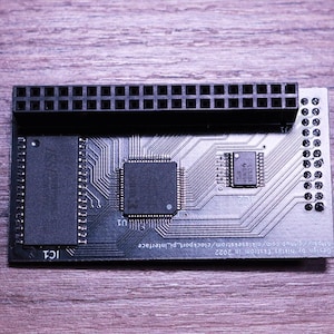 May include: A black circuit board with multiple integrated circuits, a large connector, and intricate wiring patterns. The board has the text "IC1" and other small text. The background is a wooden surface.