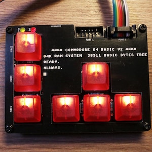 May include: A black Commodore 64 Basic V2 circuit board with red illuminated buttons. The text "64K RAM SYSTEM 38911 BASIC BYTES FREE READY. ALWAYS." is printed on the board. Wires and connectors are visible.