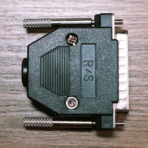 May include: A black and silver D-sub connector with the text "R/S" on a wooden surface. The connector has two silver screws and two silver posts with textured ends. The connector is used for connecting electronic devices.