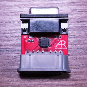 May include: A red circuit board with a black connector and a gray connector. The board has a small chip in the center and the text "ARTHRIMUS.COM" in white. The background is a wooden surface.