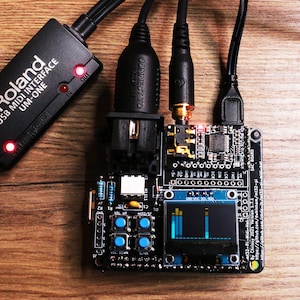 Può includere: Un'interfaccia MIDI USB Roland UM-ONE nera collegata a un circuito stampato con più cavi. La scheda ha un piccolo schermo che mostra una grafica blu e diversi pulsanti. L'installazione è su una superficie di legno.