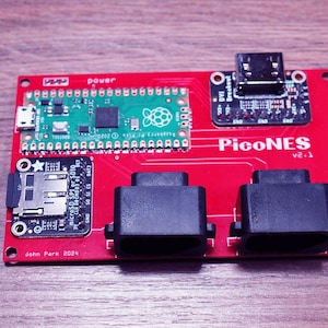 Könnte beinhalten: Eine rote PicoNES V2.1-Leiterplatte mit verschiedenen elektronischen Komponenten, darunter ein grüner Raspberry Pi Pico, schwarze Anschlüsse und ein Micro-SD-Kartensteckplatz. Die Platine trägt den Text "PicoNES" und "John Park 2024".
