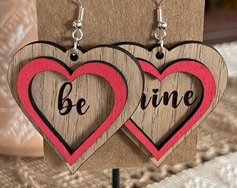 May include: Wooden heart-shaped earrings with a red outline and the words "be mine" in black script. The earrings are displayed on a brown card, perfect for Valentine's Day or a romantic gift.