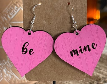 May include: Pink heart-shaped earrings with the words "be mine" in black script. The earrings are made of wood and have silver-colored hooks. A Valentine's Day themed accessory.