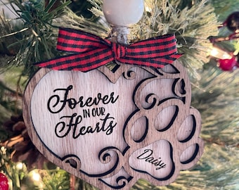 May include: Wooden heart-shaped Christmas ornament with a red and black checkered ribbon. The ornament has the text "Forever in our Hearts" and a paw print with the name "Daisy" inscribed.