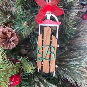 May include: Wooden sled Christmas ornament with a red bow and the letter "B". The sled has a brown wooden body, white runners, and a red accent. The ornament hangs from a natural twine string.