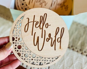 May include: A set of wooden baby milestone discs. The front disc says "Hello World" with a crescent moon design. Other discs in the box are engraved with months and numbers. The discs are made of light-colored wood.