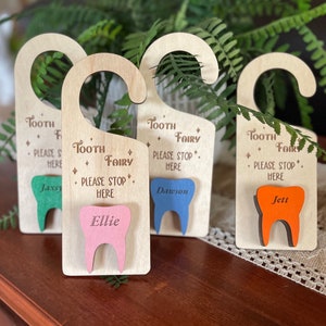 May include: Four wooden tooth fairy door hangers with a tooth cutout in different colors. The hangers have the text "Tooth Fairy" and "Please Stop Here" with a name on each tooth cutout: Jaxxy, Ellie, Dawson, and Jett.