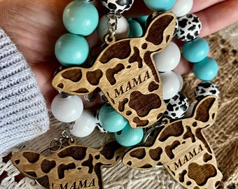 May include: A set of two wooden cow head keychains with the word "MAMA" engraved on each. The keychains are attached to a beaded bracelet with white, black and white spotted, and turquoise beads.