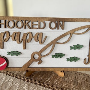May include: Wooden sign with the words "HOOKED ON papa" and a fishing rod design. The sign has a white background with brown trim and includes green fish with names. A red and white ornament is on the left.
