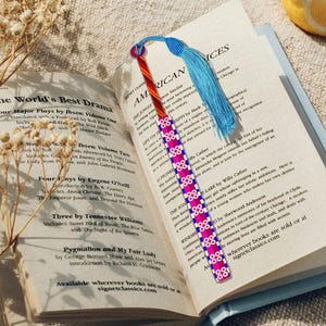 Peut inclure: Un livre ouvert avec un signet coloré. Le signet présente un motif rayé rouge, blanc et bleu avec un pompon bleu. Le livre est ouvert sur une page de texte, avec des fleurs séchées et des tranches de citron en arrière-plan.