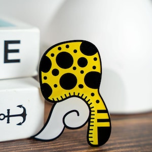 May include: A whimsical, yellow and black polka-dotted brooch with a white, swirled tail. The brooch has a ruler-like design on the side. The brooch is on a wooden surface.