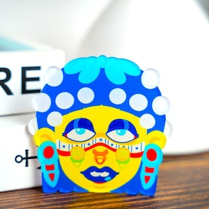 May include: A colorful, stylized face-shaped object with a blue head covering adorned with white circles. The face is yellow with blue eyes, a red nose, and blue lips. Red and blue accents frame the face. The object is on a wooden surface.