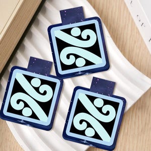May include: Three square bookmarks with a dark blue border and a light blue and black design. Each bookmark has a dark blue tab with a starry pattern. The bookmarks are on a white tray.