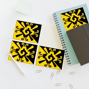 May include: Four square stickers with a black and yellow sunburst design are displayed on a white surface. A white pen, a light blue spiral notebook, a dark gray notebook, and paper clips are also visible.