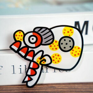 May include: A whimsical, cartoon-style fish-shaped sticker with a black outline. The design features red, white, yellow, and gray elements, including polka dots and stripes. The sticker is placed on a stack of white books.