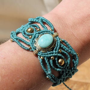 Puede incluir: Pulsera de macramé turquesa con una piedra ovalada turquesa y detalles en latón. La pulsera hecha a mano presenta intrincados nudos y pequeñas cuentas doradas, creando un accesorio de estilo bohemio.