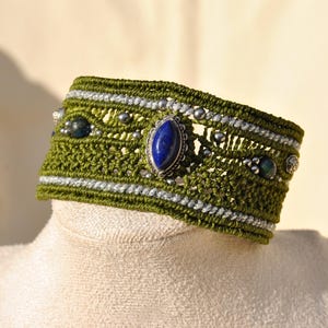May include: A handcrafted green macrame bracelet with silver accents and a large oval blue gemstone centerpiece. The bracelet also features smaller blue and silver beads. This unique piece is a statement accessory.