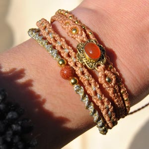 Pode incluir: Uma coleção de pulseiras de macramé em tons de bege, dourado e prateado. Uma pulseira apresenta uma pedra preciosa laranja oval em uma moldura dourada. Outras pulseiras incluem contas douradas e um cordão prateado torcido.