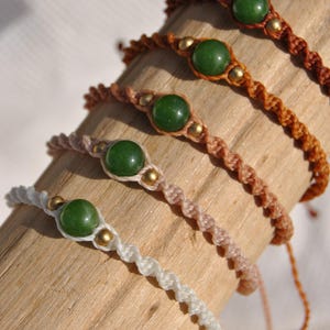 May include: Five macrame bracelets, each featuring a central green stone and gold-coloured beads. The bracelets are in white, pink, and brown hues, with a twisted rope design. They are displayed on a light brown wooden surface.