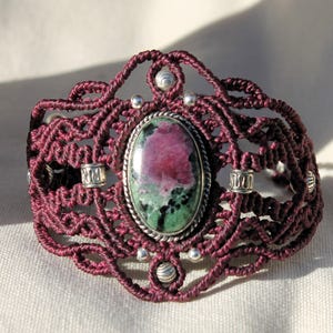 May include: A maroon macrame bracelet featuring a green and pink gemstone set in a silver bezel. The bracelet is adorned with silver beads.