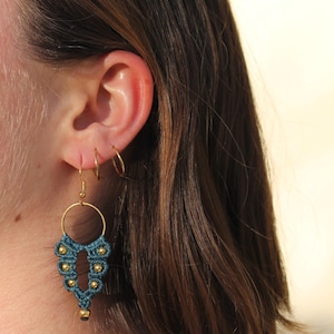 Puede incluir: Pendientes de macramé azul turquesa con cuentas doradas y un aro dorado. Los pendientes cuelgan de la oreja de una persona.