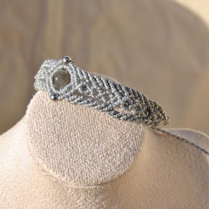 May include: A handmade macrame bracelet in shades of gray, featuring a central gray gemstone and small silver beads. The bracelet is displayed on a soft, off-white surface, showcasing its intricate woven design.