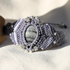 May include: A gray macrame bracelet with a white and gray stone in the center. The bracelet is decorated with silver beads and flower-shaped charms.