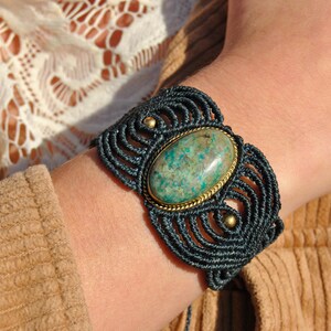 Puede incluir: Una pulsera de macramé azul oscuro con una piedra ovalada de color turquesa engastada en un marco de metal dorado. La pulsera se lleva en la muñeca, con una chaqueta de pana marrón y una blusa de encaje blanco en el fondo.